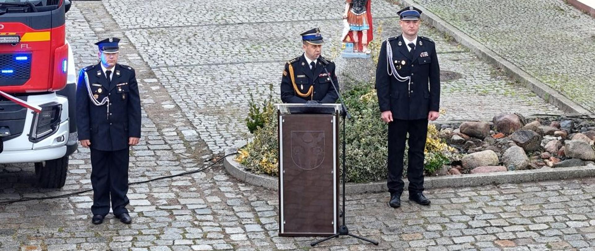 Na zdjęciu widoczny jest nowoczesny samochód ratowniczo-gaśniczy marki Volvo, przygotowany do uroczystego przekazania jednostce OSP Karlino. Pojazd udekorowano biało-czerwoną wstęgą, symbolizującą jego oficjalne wprowadzenie do służby. Przed wozem stoją druhowie w umundurowaniu galowym, a przy mównicy odbywają się przemówienia gości. W tle widoczne są pozostałe pojazdy pożarnicze jednostki. Sceneria podkreśla uroczysty i podniosły charakter wydarzenia.