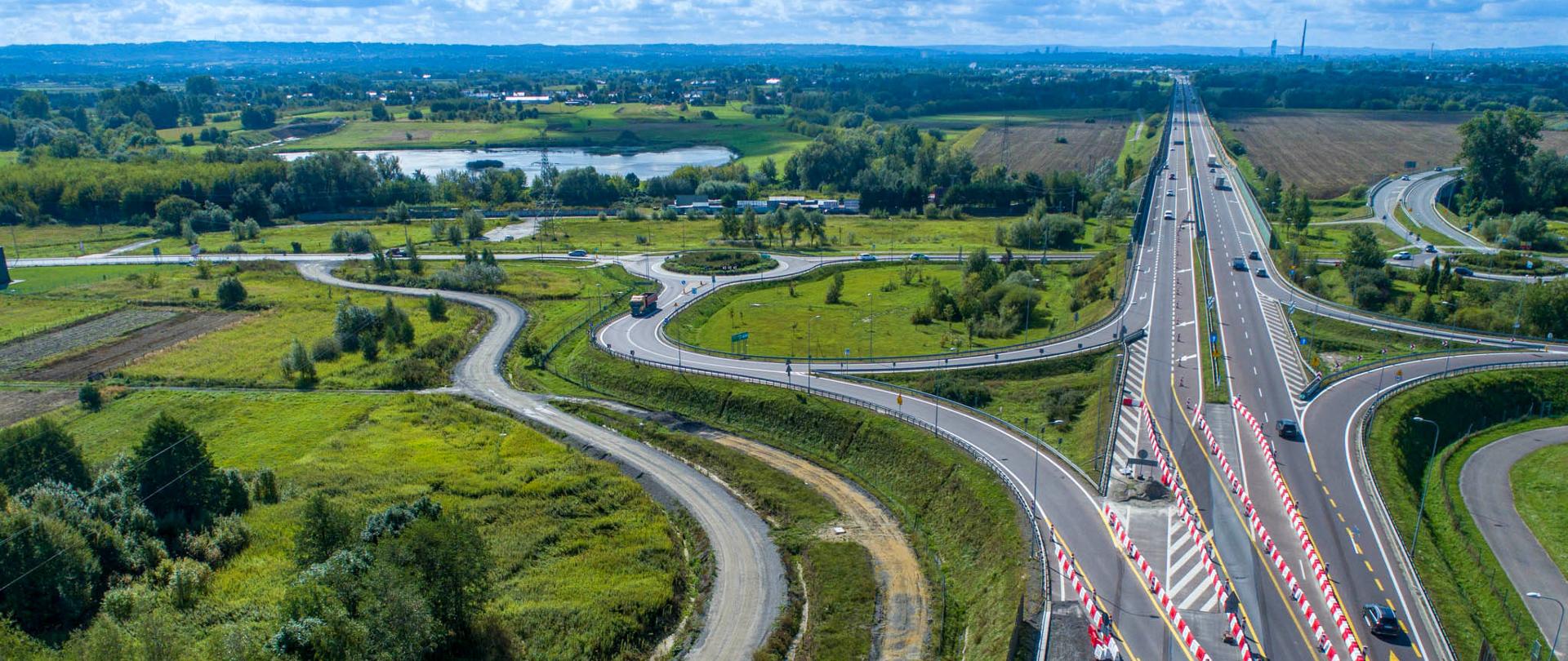 Zdjęcie przedstawia widok z lotu ptaka na rozbudowane skrzyżowanie dróg ekspresowych w otoczeniu zielonych pól i lasów. Na środku obrazu biegnie szeroka, wielopasmowa droga z wyznaczonymi pasami ruchu i barierami z biało-czerwonymi oznaczeniami, wskazującymi na trwające prace budowlane. Po obu stronach głównej trasy znajdują się zakręcające łącznice, częściowo utwardzone, a w tle widać rozległe pola, zbiornik wodny 