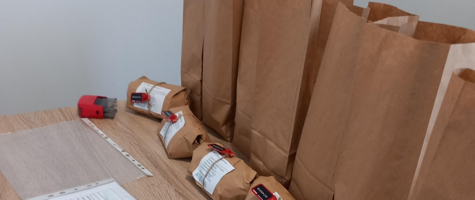 Na stole leżą zaplombowane papierowe paczki z próbkami oraz dokumenty, obok stoją większe papierowe torby przygotowane do pakowania