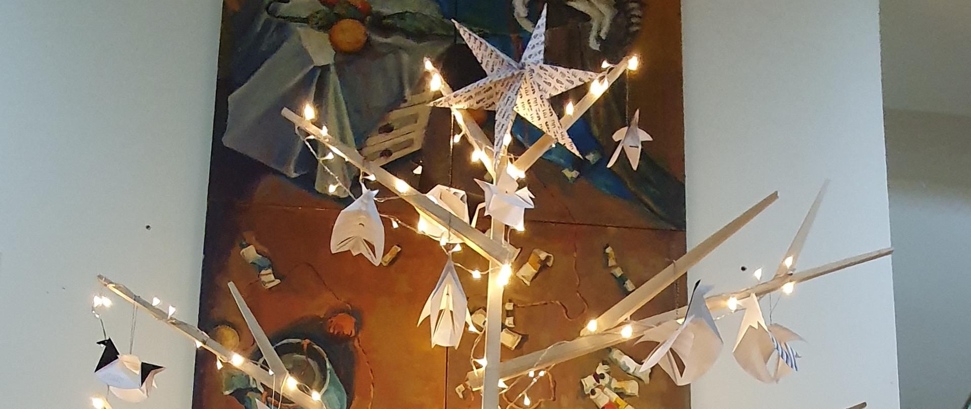 Na zdjęciu w części centralnej znajduje się choinka wykonana przez uczniów i nauczycieli PLSP im. A. Kenara w Zakopanem. Choinka wykonana jest z drewnianych listew, które są ze sobą połączone tworząc kształt drzewka. Cała konstrukcja jest oświetlona lampkami ledowymi. Na szczycie choinki zawieszona jest gwiazda wykonana z papieru. Ozdoby choinkowe wykonane z papieru, to pegazy symbolizujące logo szkoły. Pod choinką na drewnianej podłodze umieszczone są pudełka ze świątecznymi prezentami. Na drugim planie znajduje się ściana, do połowy drewniana. W górnej części zdjęcia znajduje się obraz olejny zrealizowany przez Katarzynę Jeneralczyk - uczennicę PLSP w Zakopanem 