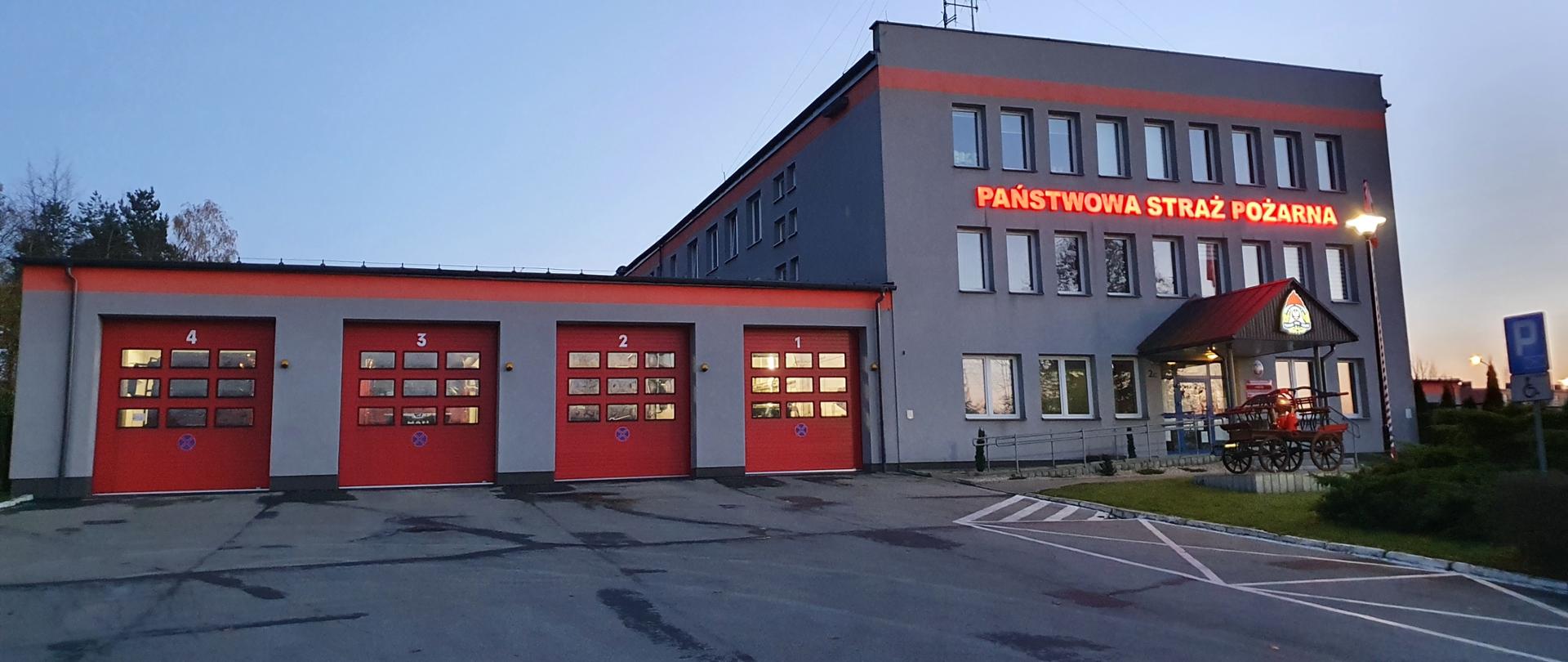 Budynek KP PSP w Olkuszu po zachodzie słońca, 4 bramy wyjazdowe wewnątrz widać pojazdy straży pożarnej, część główna budynku w kolorze szarym, budynek 3 kondygnacyjny z napisem świetlnym PAŃSTWOWA STRAŻ POŻARNA umieszczonym między 1 a 2 piętrem, przed budynkiem zabytkowa bryczka.
