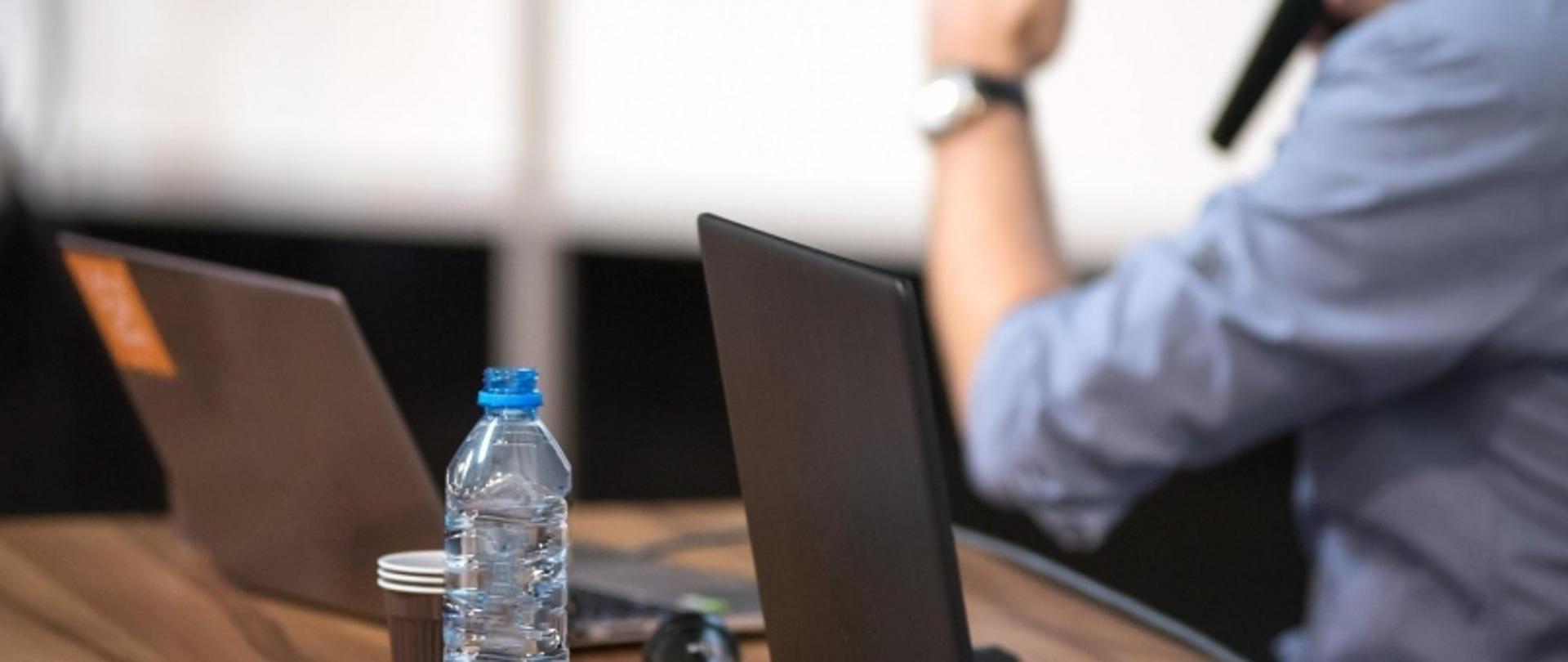 Prelegent przy stole z laptopami, mikrofonem, wodą i kawą – w trakcie wystąpienia.