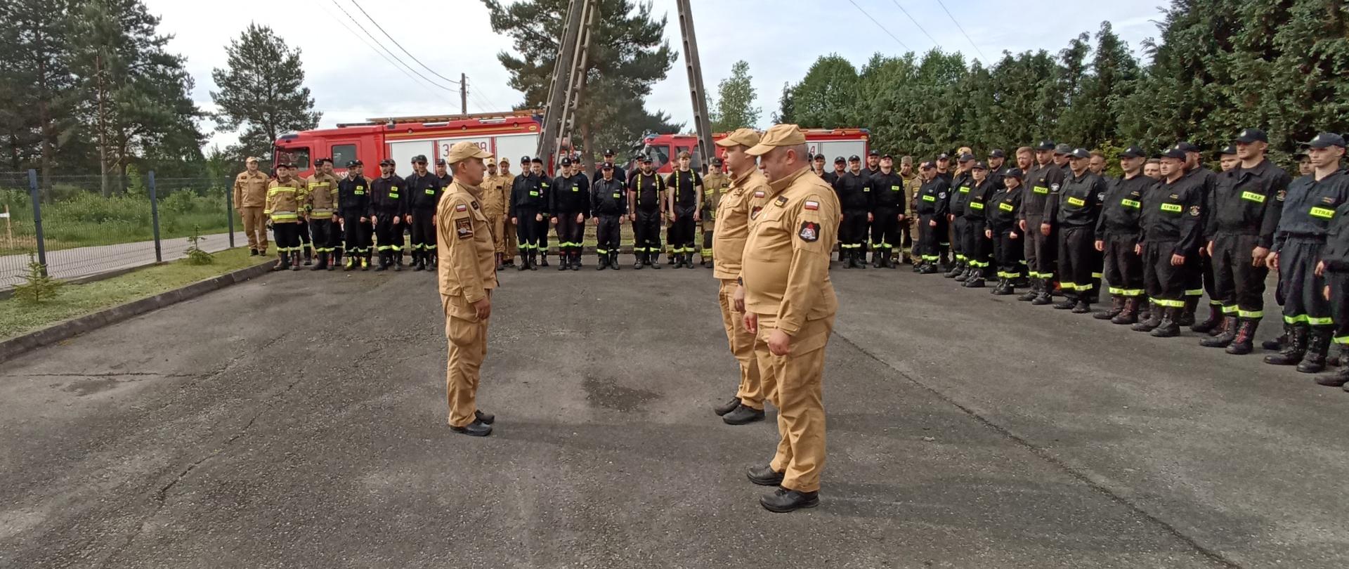 Na zdjęciu, na zewnątrz duża grupa strażaków stojących na utwardzonym placu. Większość strażaków jest ubrana w ciemne umundurowanie, a trzech z nich, stojących na pierwszym planie, nosi jasnobrązowy mundur. W tle widoczny jest duży, czerwony wóz strażacki oraz drzewa. Niebo jest pochmurne, ale jasne.