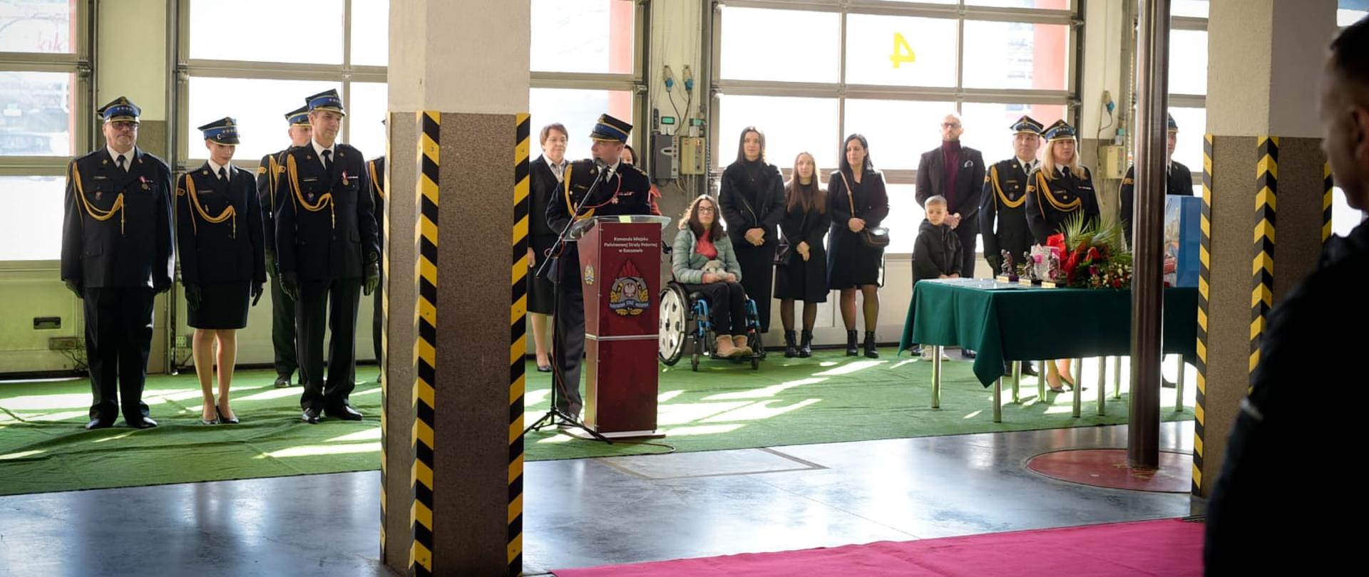 Zdjęcie przedstawia strażaków odchodzących na zaopatrzenie emerytalne, przemawiającego komendanta miejskiego PSP w Rzeszowie oraz przybyłych gości. Na zdjęciu widoczny jest stolik z pamiątkami dla odchodzących funkcjonariuszy. 