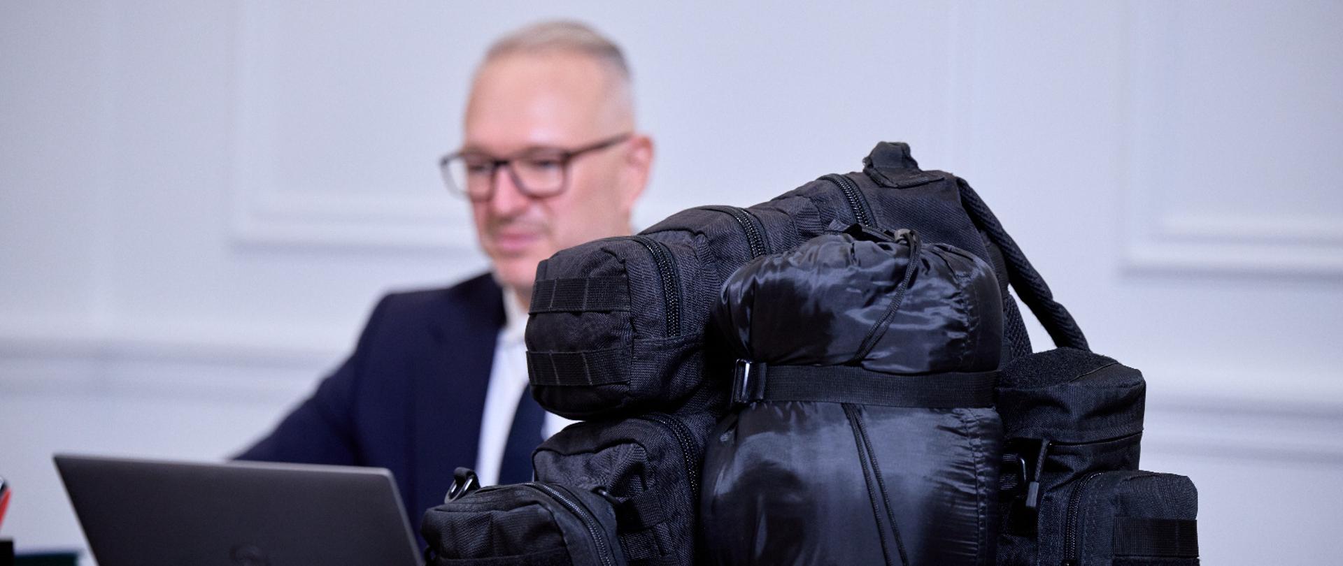 na pierwszym planie czarny plecak który leży na biurku, w tle widać zamazaną postać 