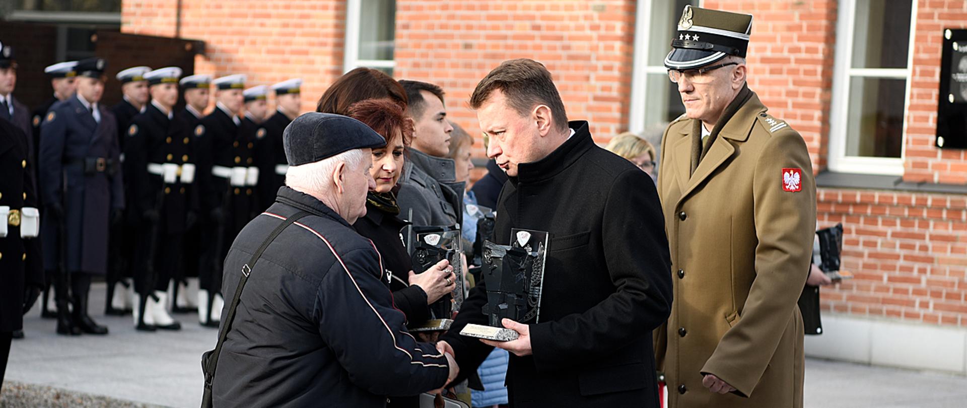 Szef MON wręcza rodzinie poległego weterana pamiątki. W tle dowódca Operacyjny Rodzajów Sił Zbrojnych i żołnierze Pułku Reprezentacyjnego.