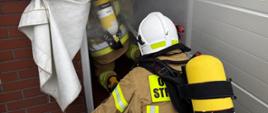 Zdjęcie przedstawia rotę strażaków w ubraniu specjalnym i SOUO oraz linią gaśniczą podczas wejścia do zadymionego pomieszczenia warsztatu JRG w Oleśnie.