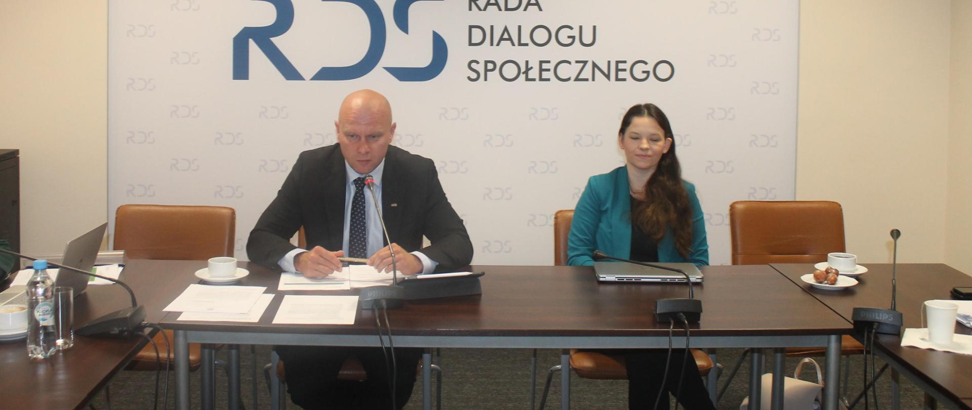 Na zdjęciu widnieje stół prezydialny za którym siedzą od lewej strony: Przewodniczący zespołu ds. usług społecznych – Bartłomiej Mickiewicz oraz Katarzyna Nowakowska - Podsekretarz Stanu w Ministerstwie Rodziny, Pracy i Polityki Społecznej. W tle siedzących znajduje się baner z napisem Rada Dialogu Społecznego a nad nim wisi godło Polski.
