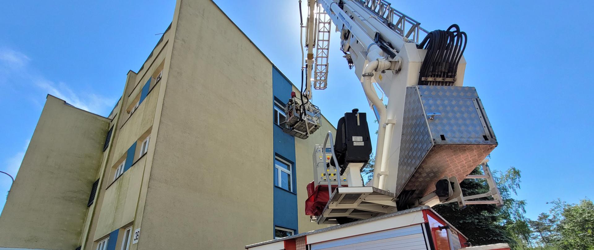 Tylna część podnośnika strażackiego z rozłożonym koszem sięgającym do okna na drugim piętrze budynku biurowego. 