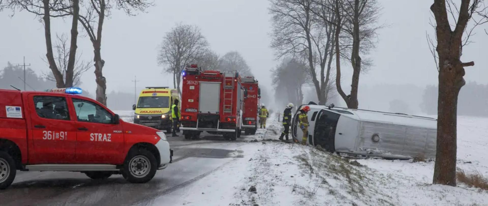 Cztery pojazdy znajdujące się na drodze. Trzy pożarnicze i jedna karetka pogotowia. Po prawej stronie jezdni przewrócony na boku samochód bus.