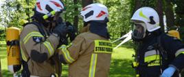 Ćwiczenia w Pakosławiu. Na tle parku stoi trzech strażaków. Dwóch z nich ma na sobie ubrania w kolorze piaskowym, trzeci - granatowym. Strażacy ubierają aparaty powietrzne.