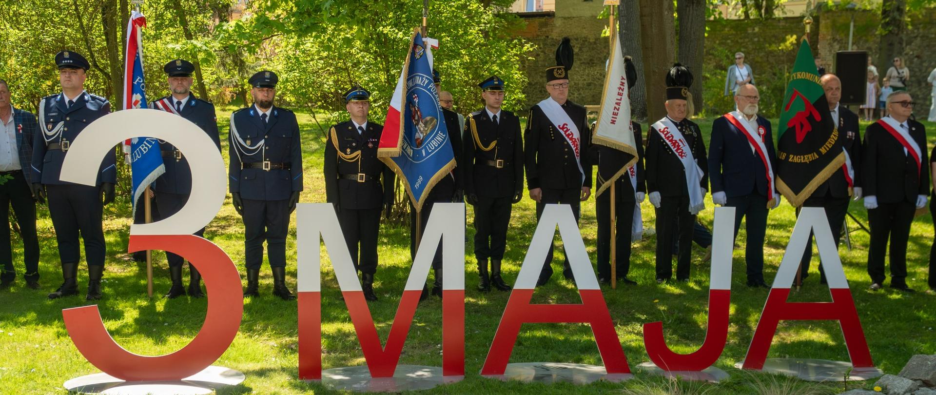 Fotografia przedstawia oficjalne uroczystości patriotyczne z okazji Święta Narodowego Trzeciego Maja, odbywające się w parku na tle zielonych drzew. Na pierwszym planie widnieje napis biało czerwony 3 maja, za nim ustawione są poczty sztandarowe.