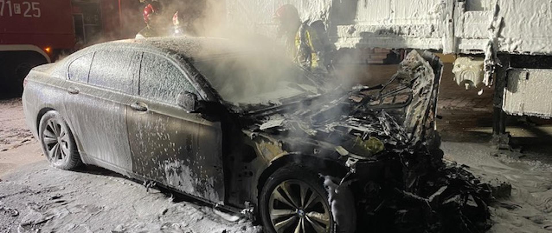 Zdjęcie przedstawia zniszczony przez pożar pojazd osobowy marki BMW. Spalona jest komora silnika obok stoi naczepa samochodu ciężarowego częściowo spalona. Oba pojazdy oblane pianą gaśniczą. W tle samochód pożarniczy oraz pracujący strażacy z JRG Międzyrzecz
