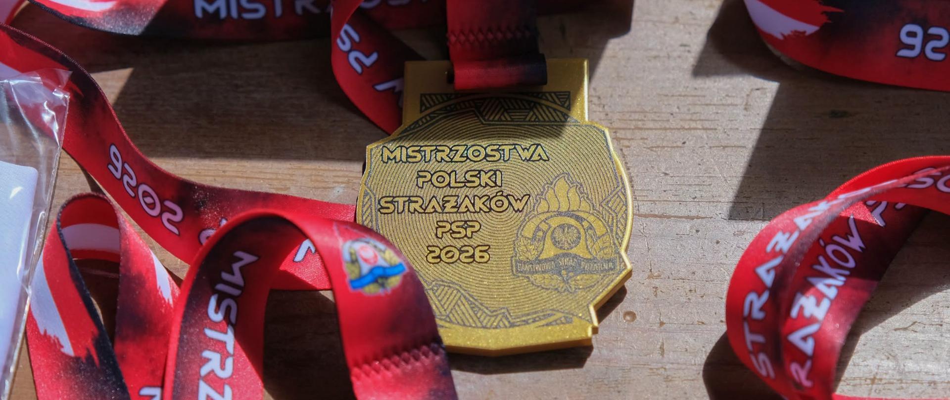 Złoty medal na czerwono-czarnej smyczy z napisem „Mistrzostwa Polski Strażaków PSP 2026”, leżący na drewnianej powierzchni.