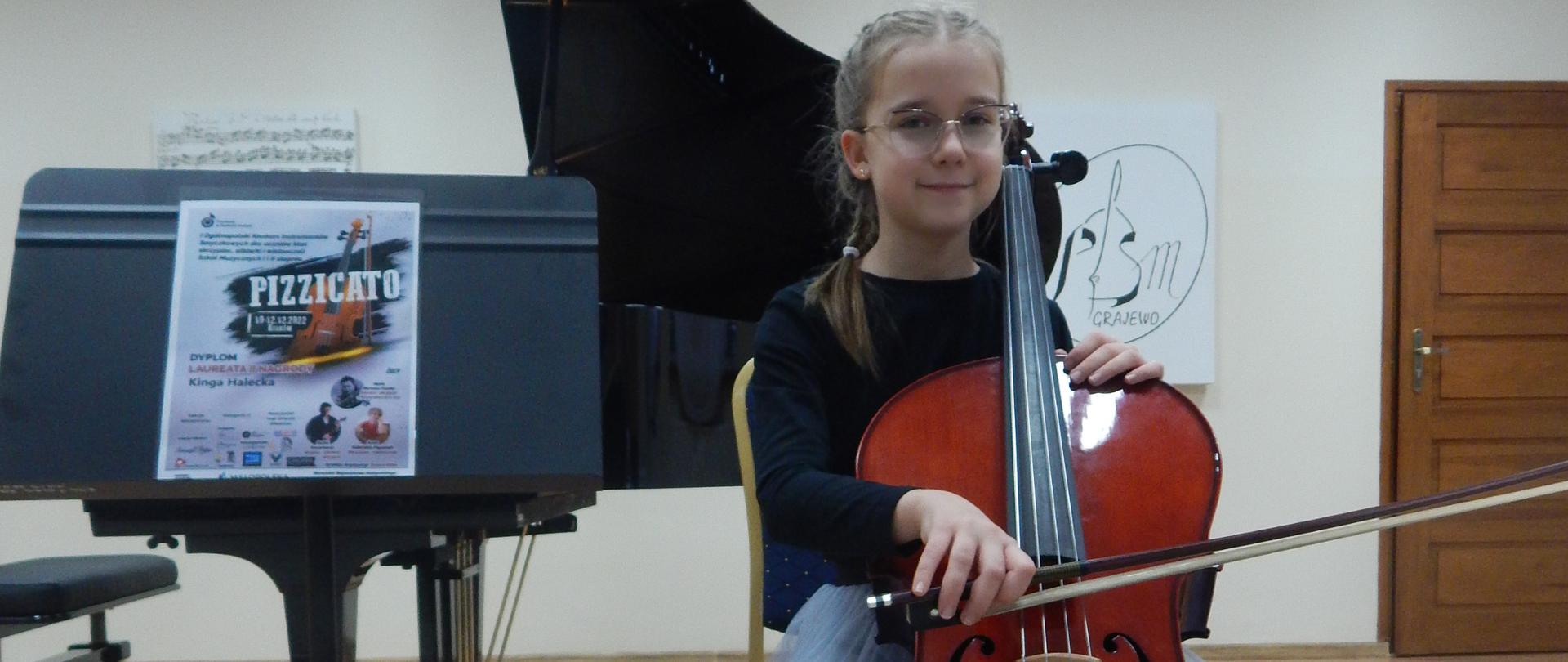 Dziewczynka ubrana w czarną bluzkę i szarą spódnicę siedzi przy wiolonczeli, obok stoi pulpit z dyplomem za zdobycie II nagrody w kategorii II w I Ogólnopolskim Konkursie Instrumentów Smyczkowych dla uczniów klas skrzypiec, altówki i wiolonczeli Szkół Muzycznych I i II stopnia "PIZZICATO" w Krakowie (edycja on-line). W tle fortepian i obrazy.