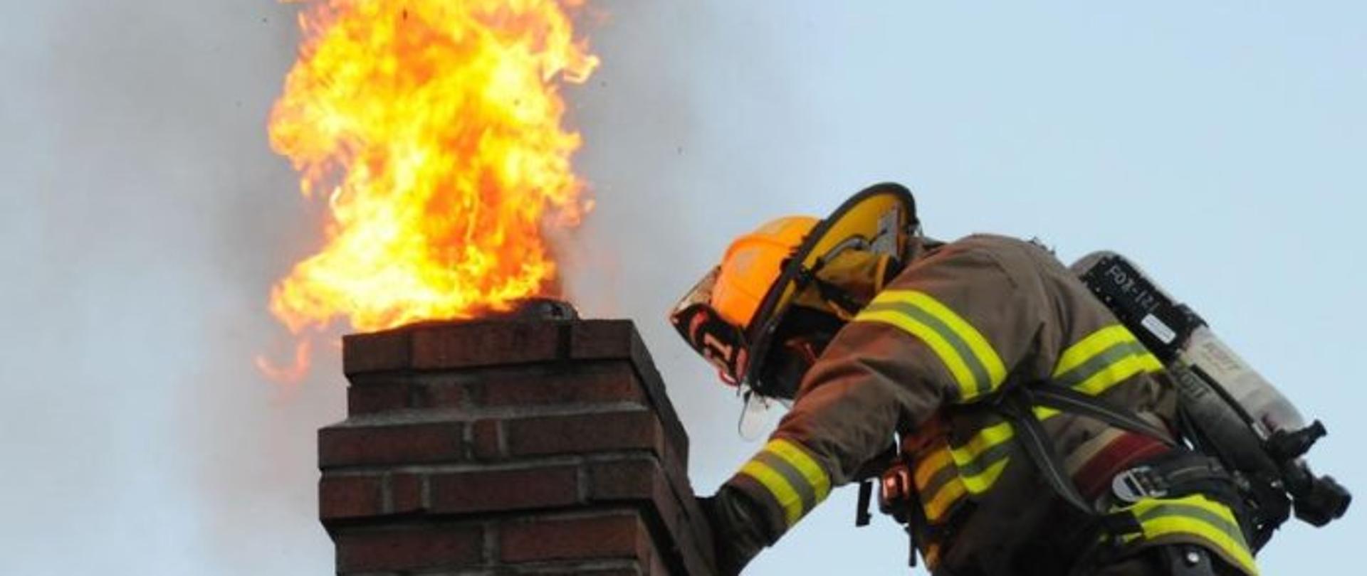 Na zdjęciu strażak w ubraniu bojowym i hełmie przy palącym się kominie z cegieł..