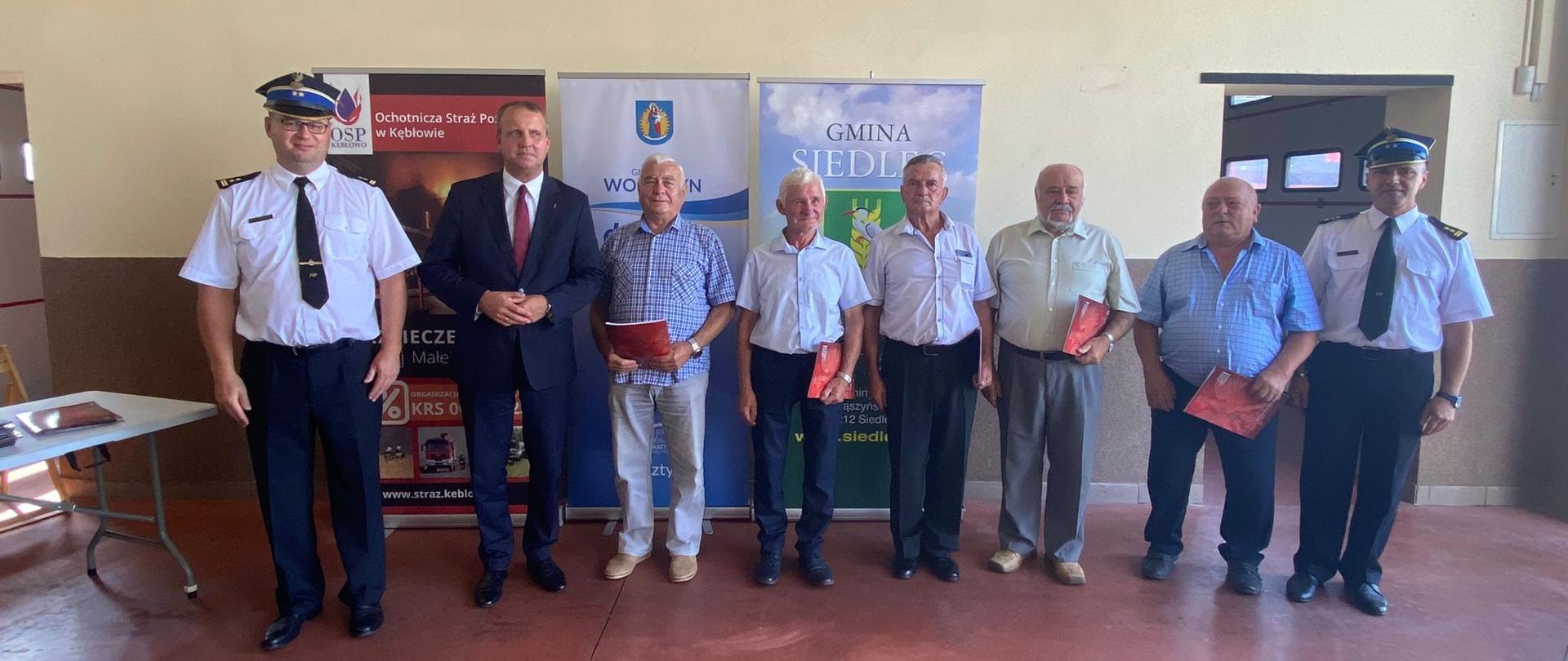 grupa mężczyzn stoi obok siebie, z lewej i prawej strony stony stoi strażak w ciemnych spodniach białej koszuli i w rogatywce