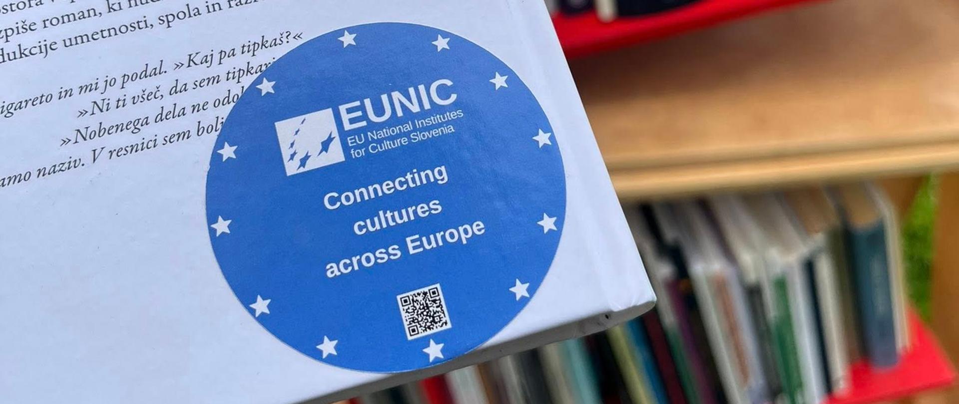 Zbliżenie na książkę w języku słoweńskim oznaczoną naklejką EUNIC Slovenia „Connecting cultures across Europe”. W tle widoczny jest plenerowy regał z książkami ustawiony na trawie w parku.