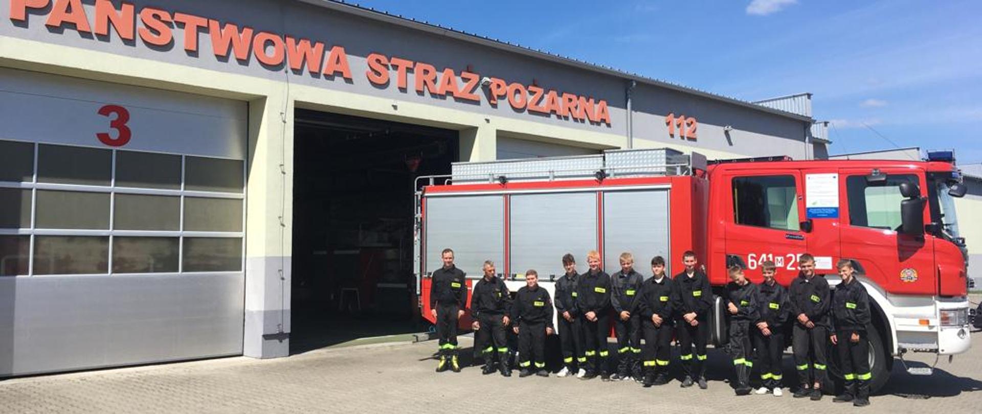 Druhowie Młodzieżowej Drużyny Pożarniczej z OSP Kosów Lacki odwiedzili KP PSP w Sokołowie Podlaskim - na zdjęciu strażacy w ubraniu koszarowym na placu przed strażnicą na zbiórce w szeregu przed czerwonym samochodem strażackim.