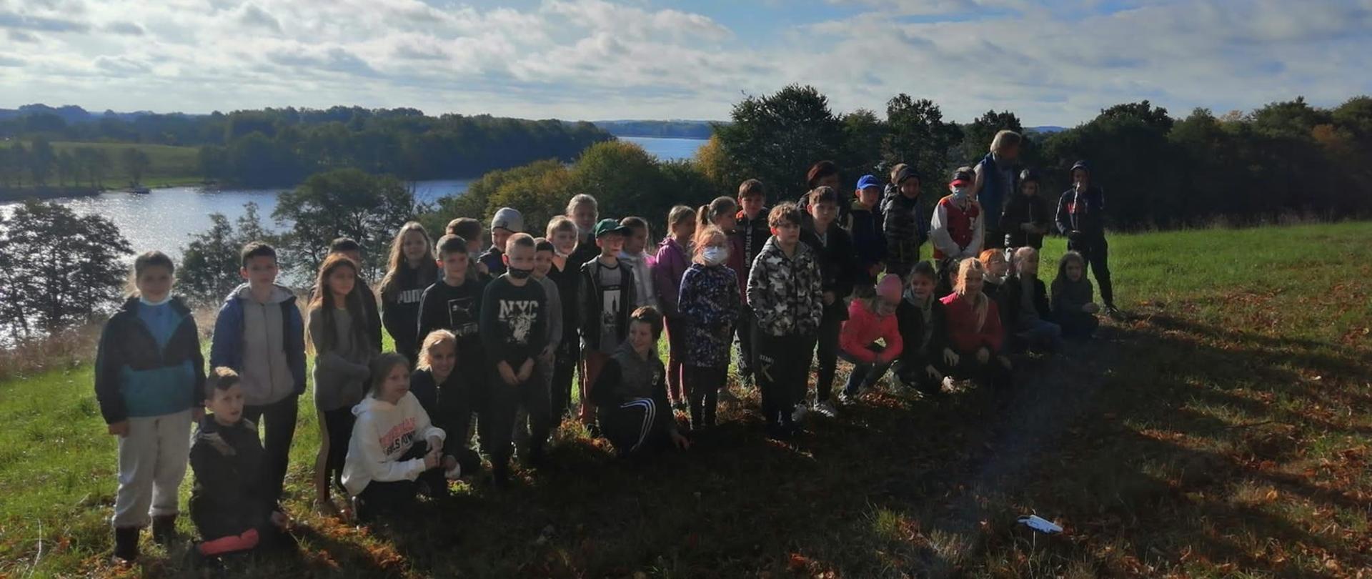 Grupa dzieci stojąca na trawie na tle rzeki i drzew.