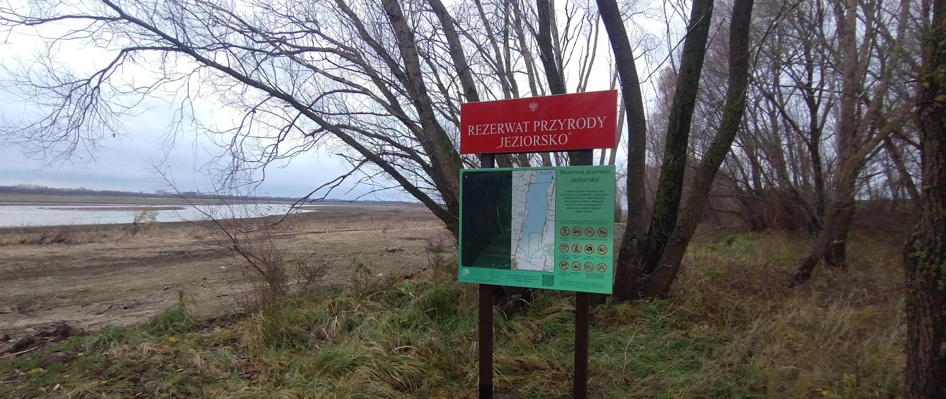 Na pierwszym planie czerwona tablica z napisem Rezerwat przyrody ,,Jeziorsko" i z godłem. Pod nią zielona tablica, na której znajduje się mapa terenu oraz oraz Napis Rezerwat przyrody Jeziorsko, a pod nim drobny druk oraz grafika. Za tablicą widoczne drzewa oraz woda.