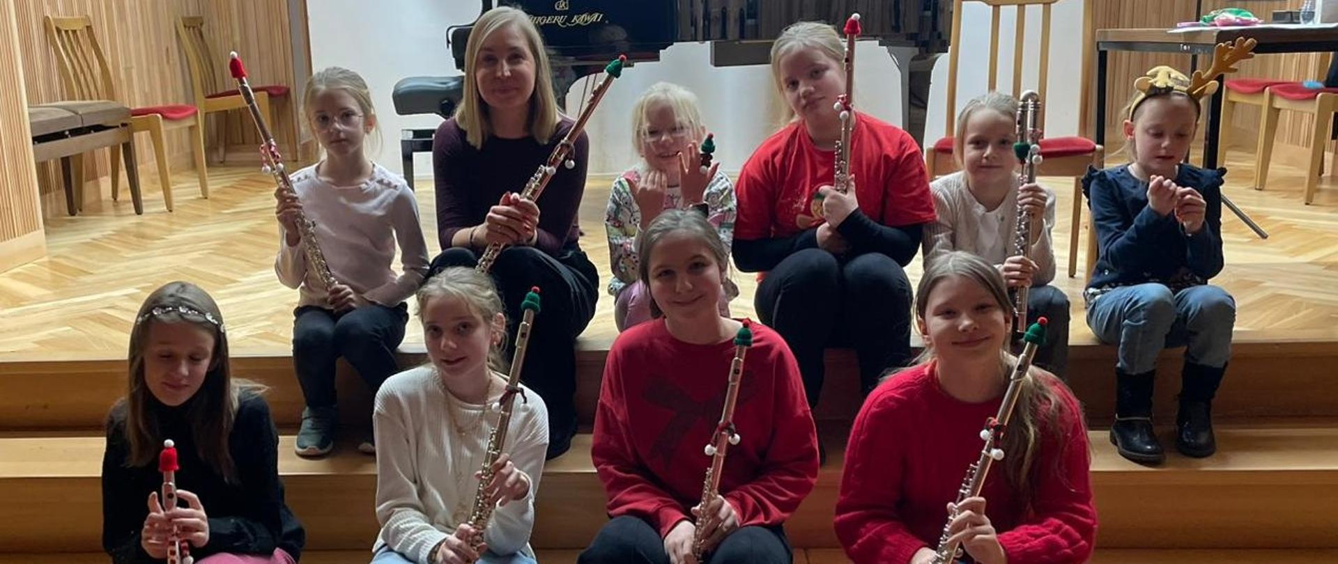 Dr Joanna Kowalczyk i dziewięć uczestniczek warsztatów siedzą na stopniach sceny, pozując do wspólnego pamiątkowego zdjęcia. Każda z osób trzyma swój flet poprzeczny pionowo, a instrumenty ozdobione są świątecznymi akcentami.