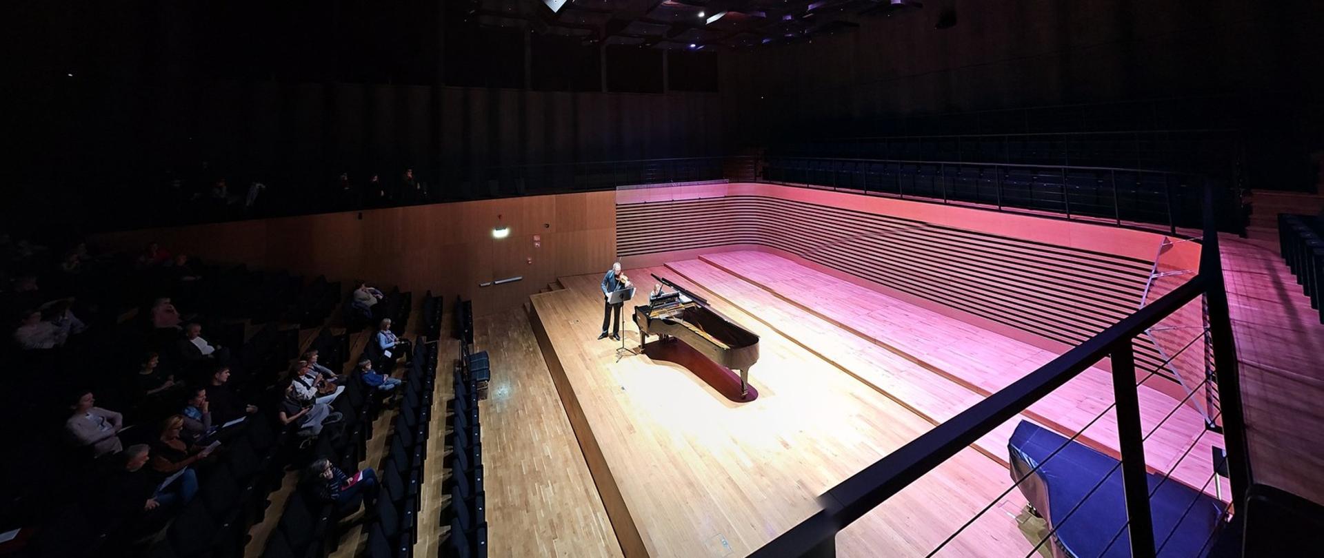 Widok z góry, z prawej strony - na środku estrady sali koncertowej mężczyzna stojący przy pulpicie gra na skrzypach, obok kobieta gra na fortepianie. Po lewej stronie widać publiczność siedzącą na widowni.