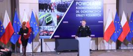 Komendant główny Państwowej Straży Pożarnej przemawia obok stoi kobieta a za nimi stoją flagi Unii Europejskiej oraz Polski telebim na którym wyświetlany jest napis powołanie koordynatorów ds. ochotniczych straży pożarnych.