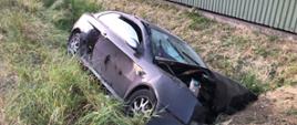 Zdjęcie przedstawia samochód osobowy koloru czarnego marki Alfa Romeo, który wypadł z drogi i zatrzymał się w przydrożnym rowie uderzając o przepust drogowy wjazdu na posesję. Samochód ma uszkodzony przód. 