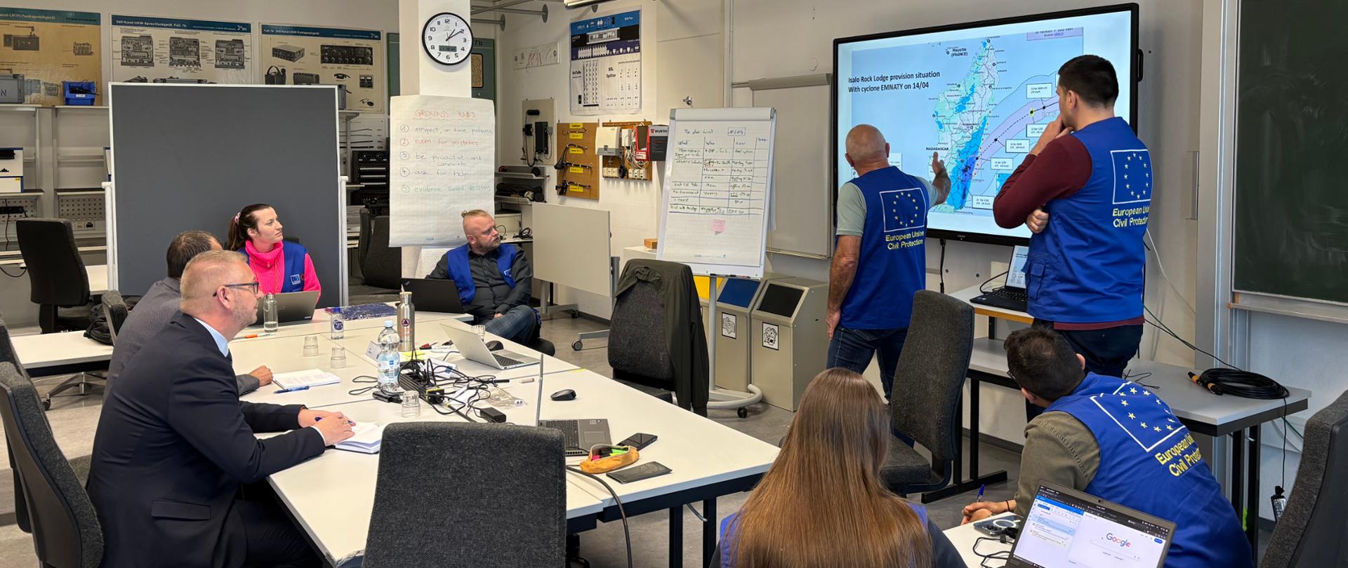 Na zdjęciu pracownicy biura unijnego mechanizmu ochrony ludności w niebieskich kamizelkach przedstawiają mapę osobą w garniturach