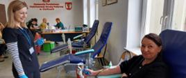Na zajęciu widoczna kobieta w czarnej koszulce i granatowych spodniach siedząca na fotelu na sali podczas oddawania krwi podczas akcji zbiórki krwi zorganizowanej przez Klub Honorowych Dawców Krwi Polskiego Czerwonego Krzyża przy Komendzie Powiatowej Państwowej Straży Pożarnej w Gorlicach w tle personel Regionalnego centrum Krwiodawstwa i Krwiolecznictwa.