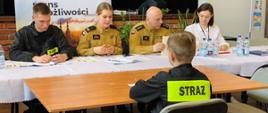 Na pierwszym planie dziecko siedzące na krześle tyłem, ubrane w ubranie koszarowe z napisem straż. Przed dzieckiem przy stole siedzi komisja czteroosobowa, składająca się z trzech strażaków w mundurach służbowych oraz jeden pracownik Gminy Przemęt. Na drugim planie baner Gminy Przemęt i czarna kotara w oddali.