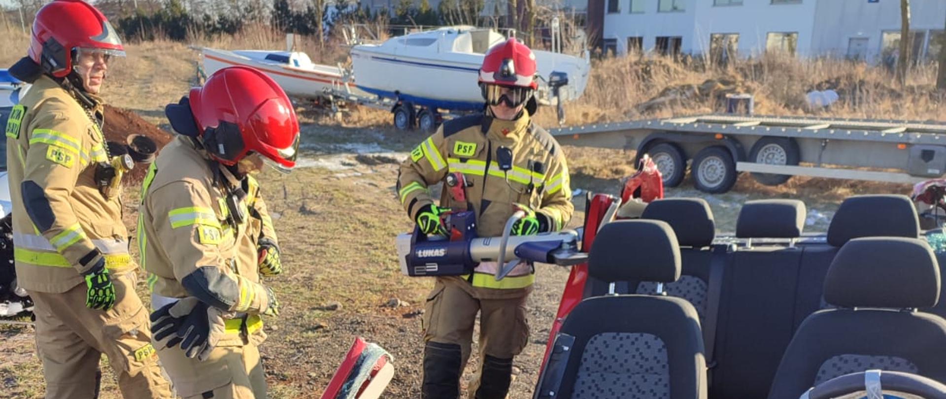 Strażacy stojący przy wraku, jeden przecina tylny słupek pojazdu, auto bez dachu