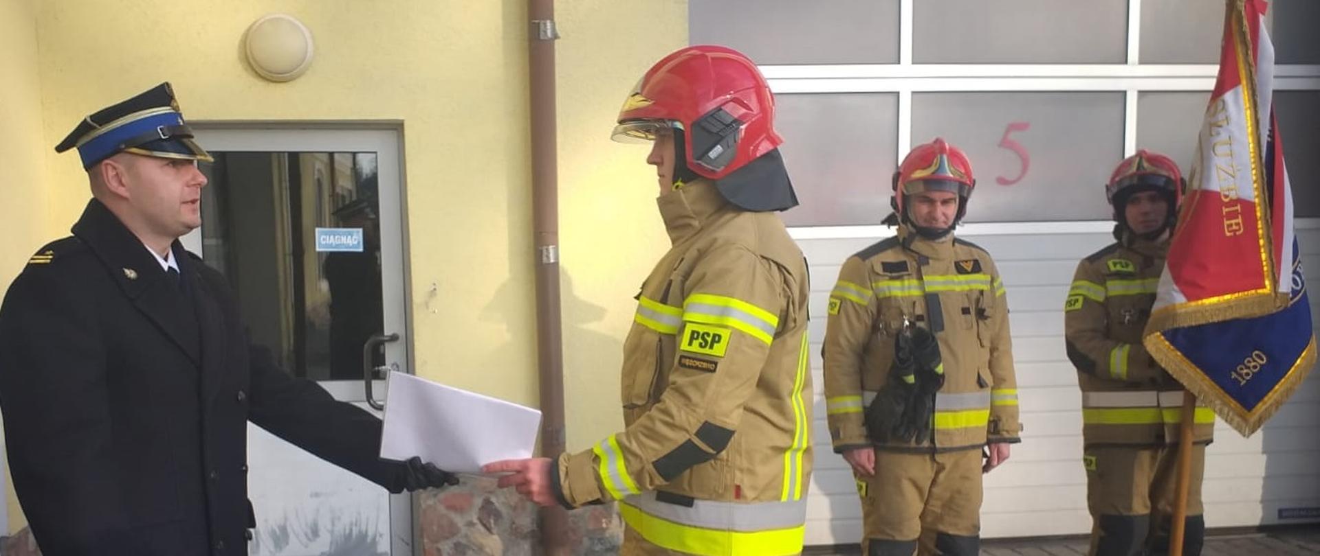 Ślubowanie strażaka. Na placu komendy strażak odbiera od Komendanta akt. Za nim widać dwóch strażaków , jedn z nich trzyma sztandar. Strażacy ubrani są w ubrania specjalne. 