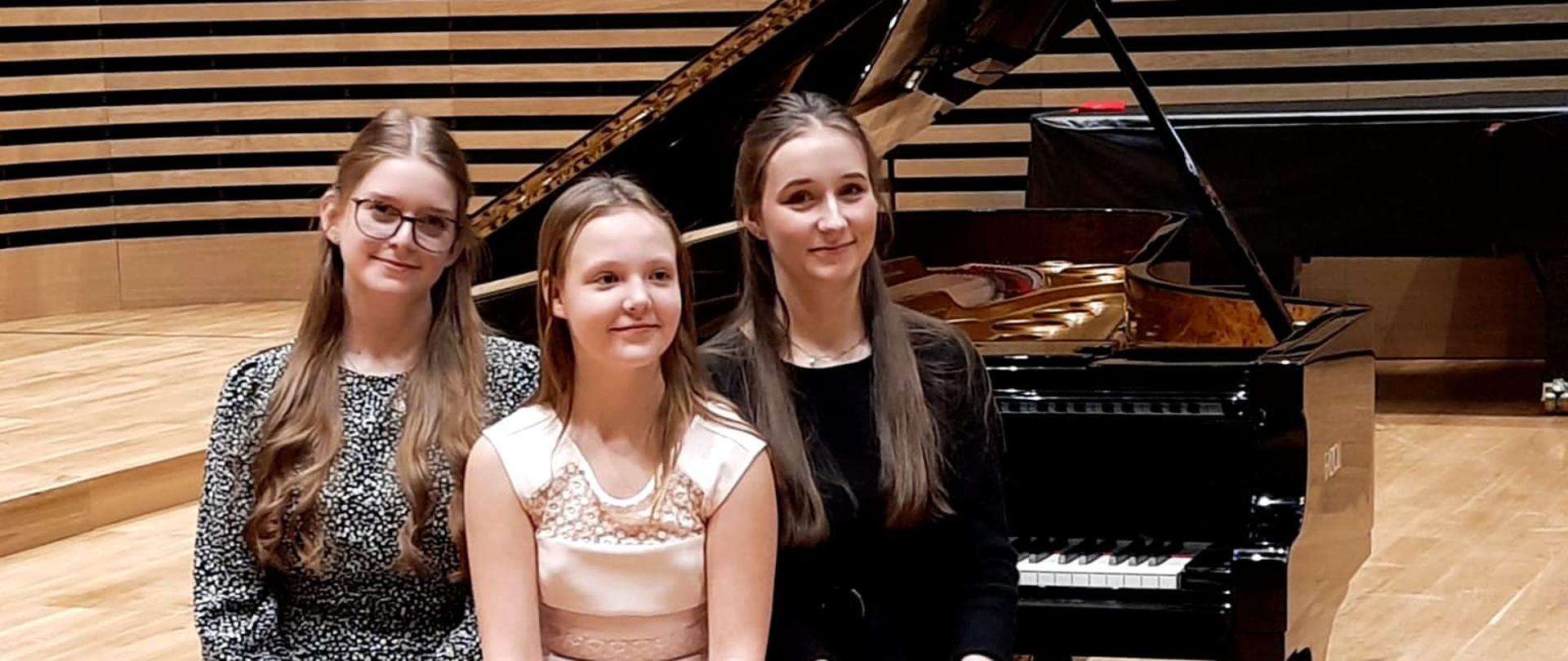 Trzy uczennice siedzące na krześle przy fortepianie na scenie sali koncertowej PSM