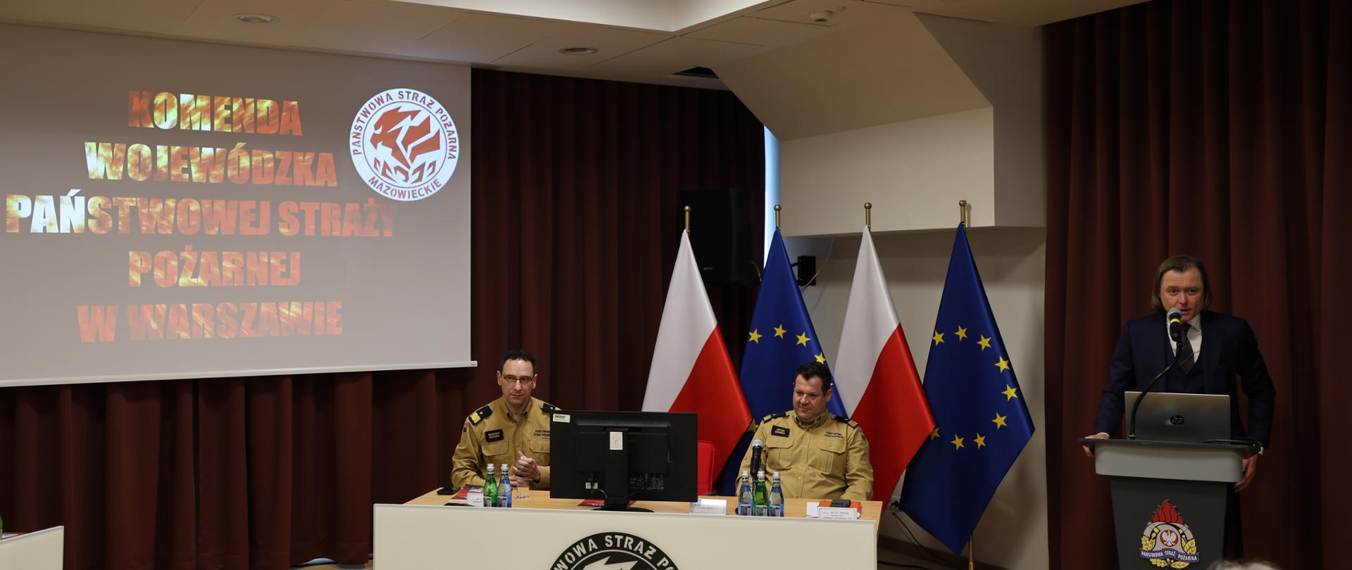 Po prawej stronie zdjęcia przy szarej mównicy z logo PSP przemawia Wojewoda Mazowiecki Mariusz Frankowski w granatowym garniturze. W centralnej części zdjęcia przy białym stole z logo KW PSP w Warszawie siedzą Komendant Główny PSP nadbryg. Wojciech Kruczek oraz Mazowiecki Komendant Wojewódzki PSP nadbryg. Artur Gonera. Za nimi widoczne flagi Polski i Unii Europejskiej. Po lewej stronie zdjęcia na ekranie projektorowym logo KW PSP w Warszawie i ognisty napis „KOMENDA WOJEWÓDZKA PAŃSTWOWEJ STRAŻY POŻARNEJ W WARSZAWIE”. Zdjęcie zrobione na jasnej sali konferencyjnej z bordowymi kotarami i czerwonymi krzesłami.