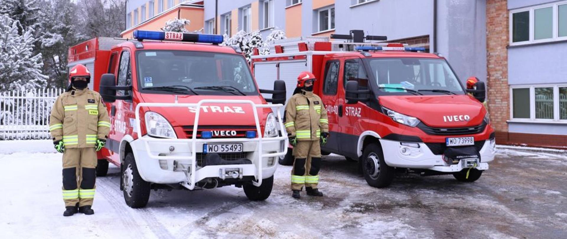 Na zdjęciu dwa samochody straży pożarnej, przy których swoi dwóch strażaków w mundurach.