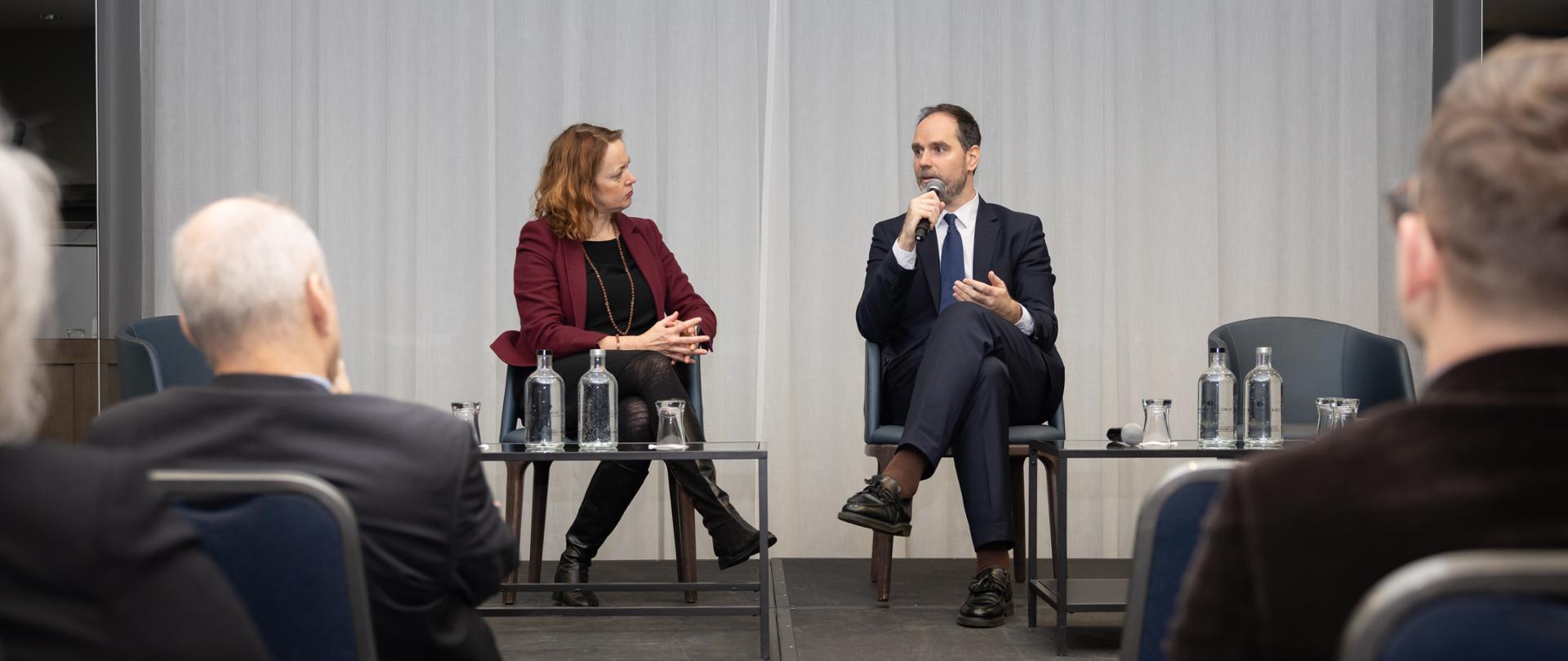 Na zdjęciu kobieta i mężczyzna siedzą na scenie. Kobieta po lewej stronie ma bordową marynarkę. Patrzy na mężczyznę po prawej, który ma na sobie ciemny garnitur i mówi do mikrofonu. Na scenie są też stoliki, na których stoją karafki z wodą i szklanki. 