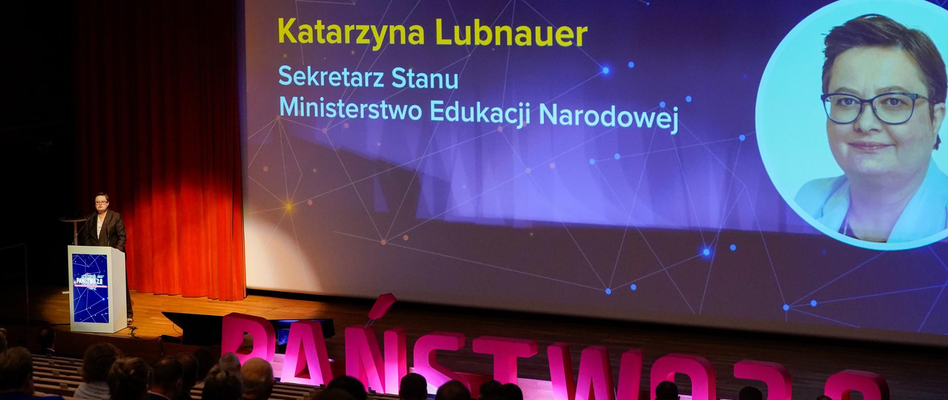 Kobieta w brązowej marynarce stoi na scenie za mównicą, przemawiając do publiczności. Na dużym ekranie w tle wyświetlany jest slajd z napisem „Proces cyfryzacji edukacji” oraz imieniem i nazwiskiem kobiety: Katarzyna Lubnauer, Sekretarz Stanu, Ministerstwo Edukacji Narodowej. Obok napisu znajduje się portret kobiety. Na pierwszym planie widać duży, różowy napis „PAŃSTWO 2.0”.