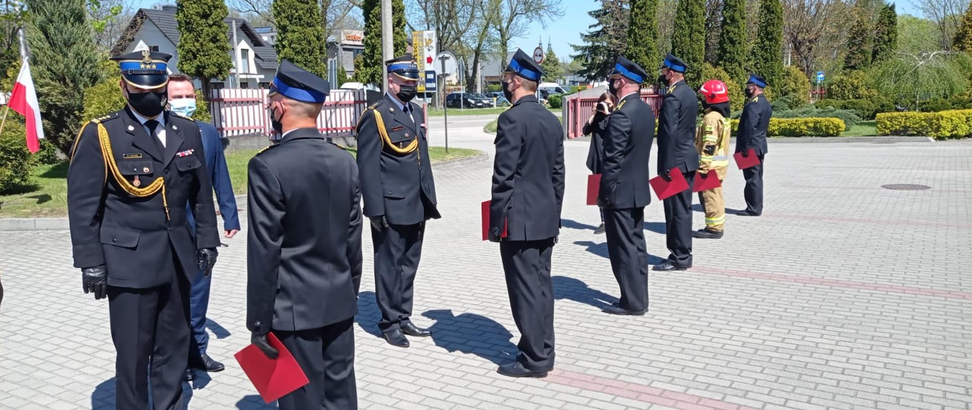 Zdjęcie przedstawia funkcjonariuszy Państwowej Straży Pożarnej w umundurowaniu galowym z założonymi maseczkami ochronnymi zakrywającymi usta i nos. Strażacy przyjmują odznaczenia i awanse od Świętokrzyskiego Komendanta Wojewódzkiego PSP. 