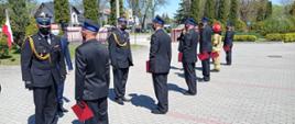Zdjęcie przedstawia funkcjonariuszy Państwowej Straży Pożarnej w umundurowaniu galowym z założonymi maseczkami ochronnymi zakrywającymi usta i nos. Strażacy przyjmują odznaczenia i awanse od Świętokrzyskiego Komendanta Wojewódzkiego PSP. 