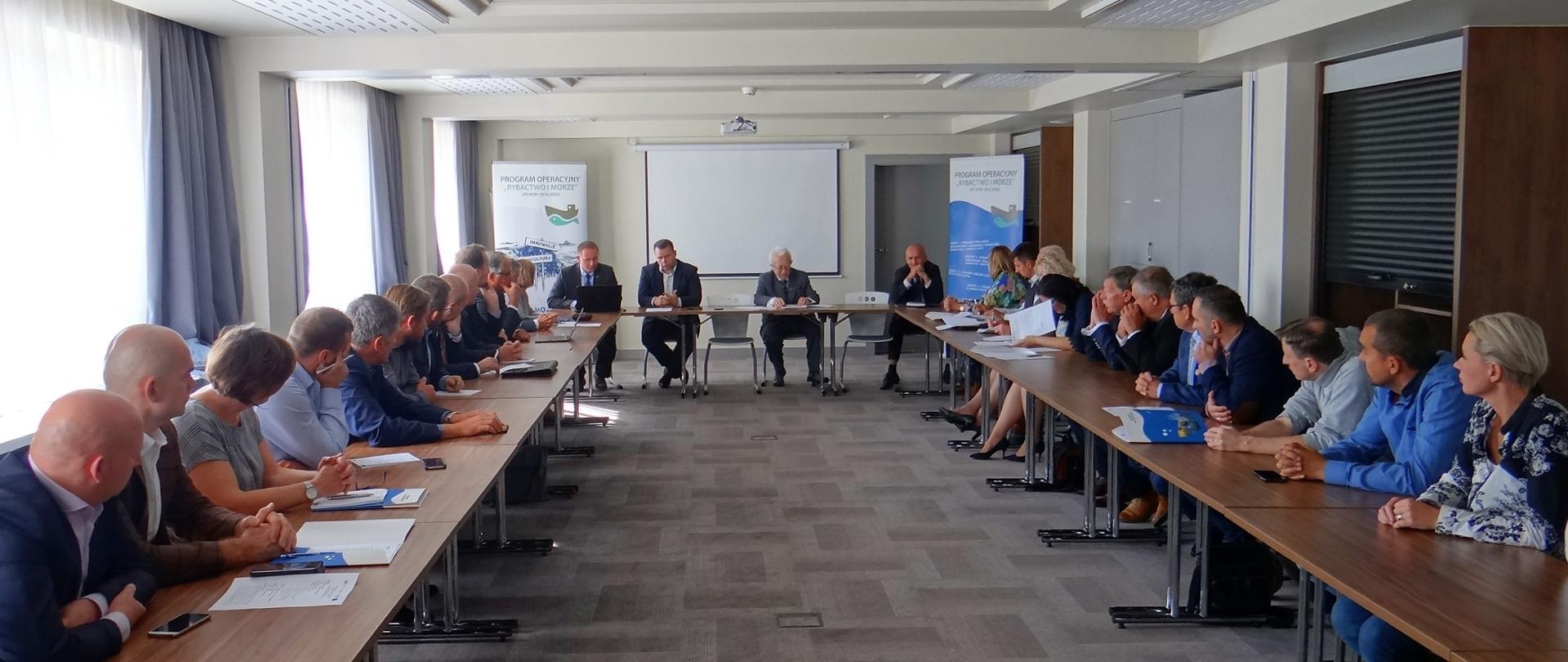 Uczestnicy posiedzenia siedzą przy wspólnym stole słuchając prelegenta