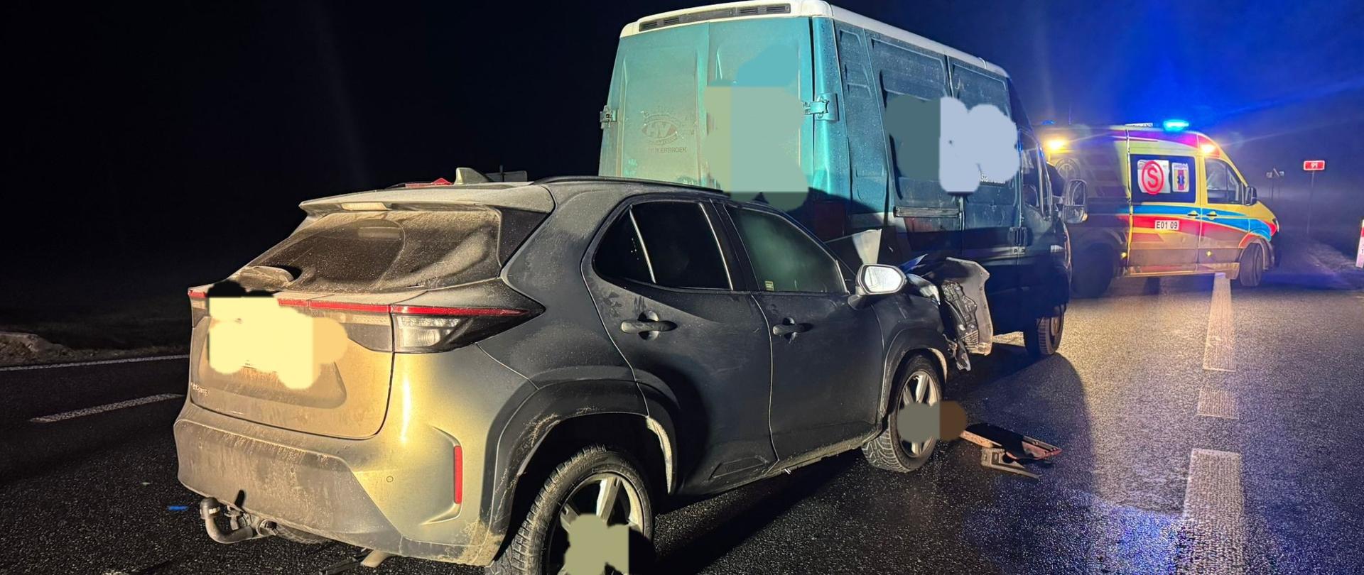 Na zdjęciu widać wypadek drogowy. Jezdnia jest mokra. Ciemny SUV stoi na prawym pasie i jest uderzony przodem w tył niebieskiego samochodu dostawczego. Przód SUV-a jest wyraźnie uszkodzony. W tle znajduje się karetka pogotowia z włączonymi sygnałami świetlnymi, co wskazuje na obecność służb ratunkowych. 