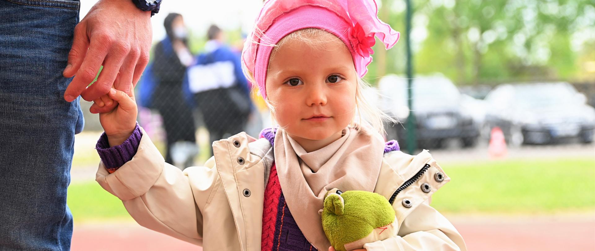 Na pierwszym planie stoi mała dziewczynka, która trzyma za rękę swojego tatę na boisku szkolnym 