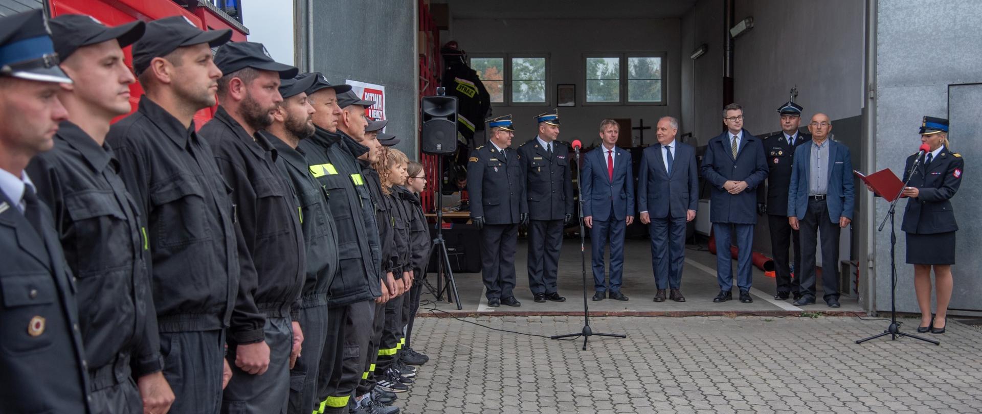 Zewnętrzy plac wyłożony kostką brukową, w tle otwarte drzwi remizy strażackiej. W lewej stronie kadru stojący w szeregu strażacy, za ich plecami widoczny fragment samochodu pożarniczego w kolorze czerwonym. W tle zdjęcia na wejściu do remizy stoją zaproszeni goście. 