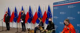 Minister Spraw Wewnętrznych i Administracji Mariusz Kamiński, Podsekretarz Stanu w MSWiA Błażej Poboży stoją przy mikrofonach, w tle flagi Polska i UE obok na krzesłach siedzą funkcjonariusze od lewej: Policji, Straży Granicznej i Państwowej Straży Pożarnej z siedzącymi obok nich psami służbowymi.