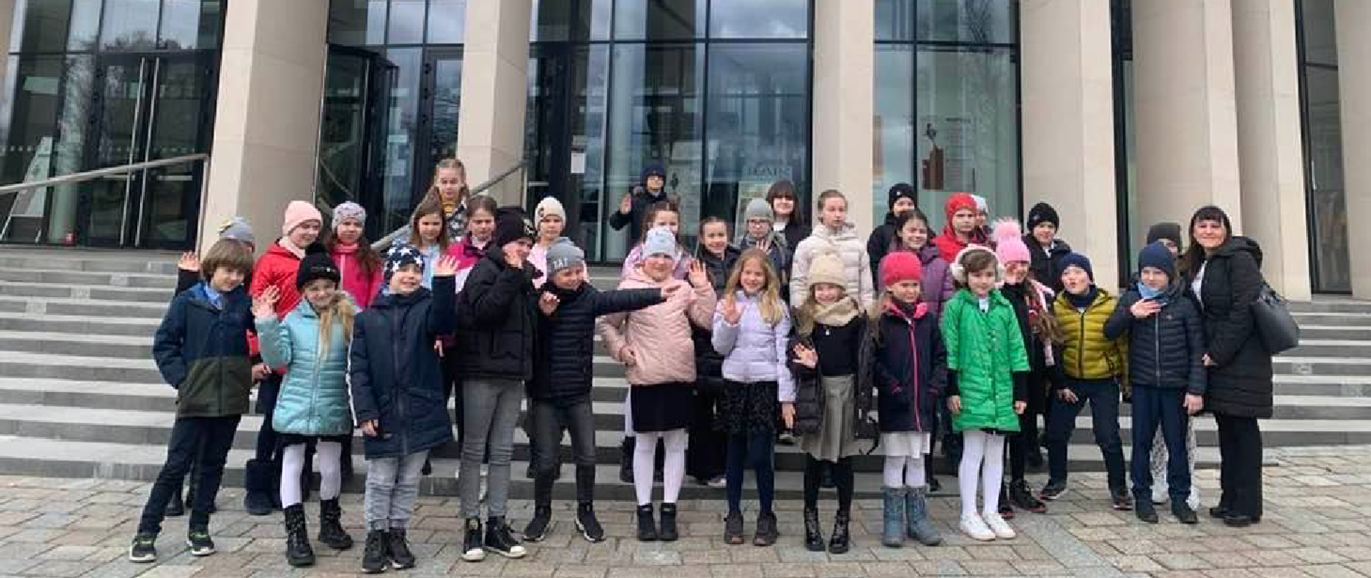 Na zdjęciu duża grupa dzieci stojąca przed schodami prowadzącymi do dużego, oszklonego budynku.