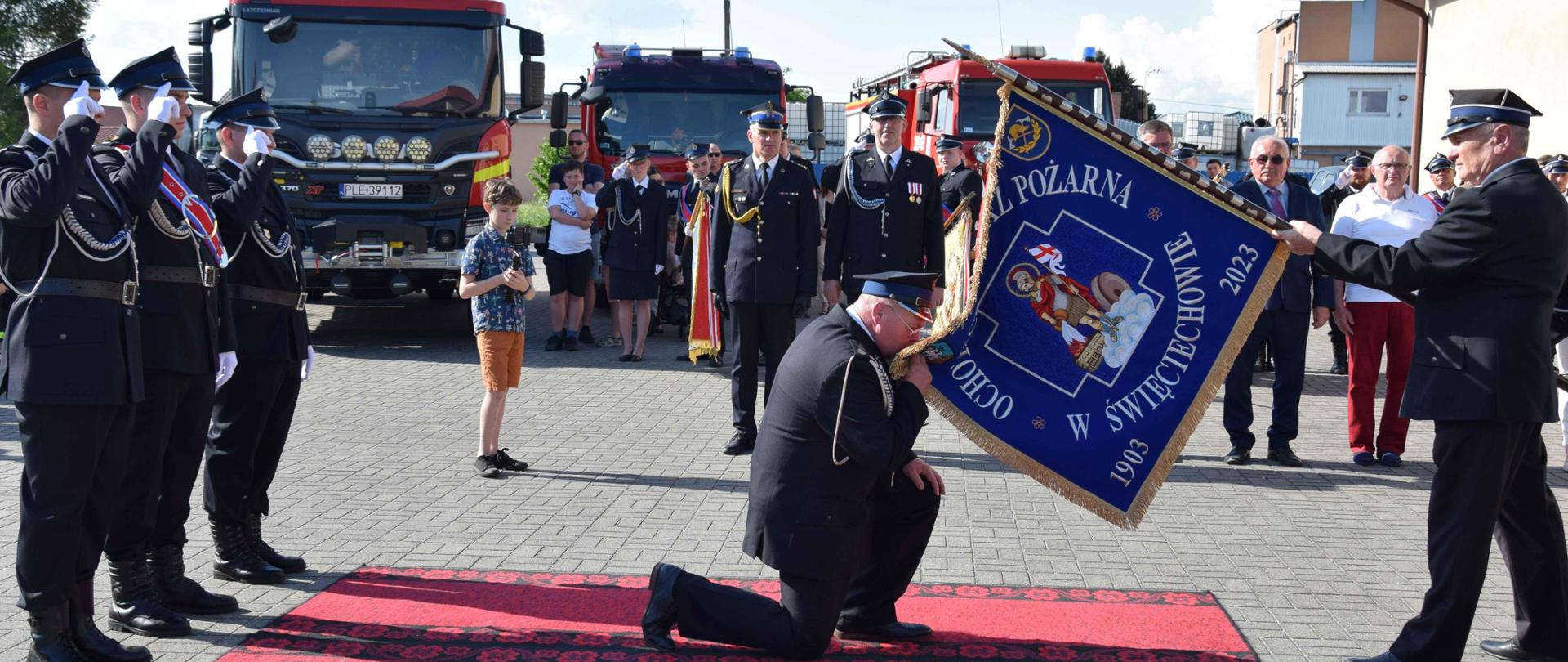 Strażacy trzymają sztandar. Drugi strażak klęczy i go całuje.