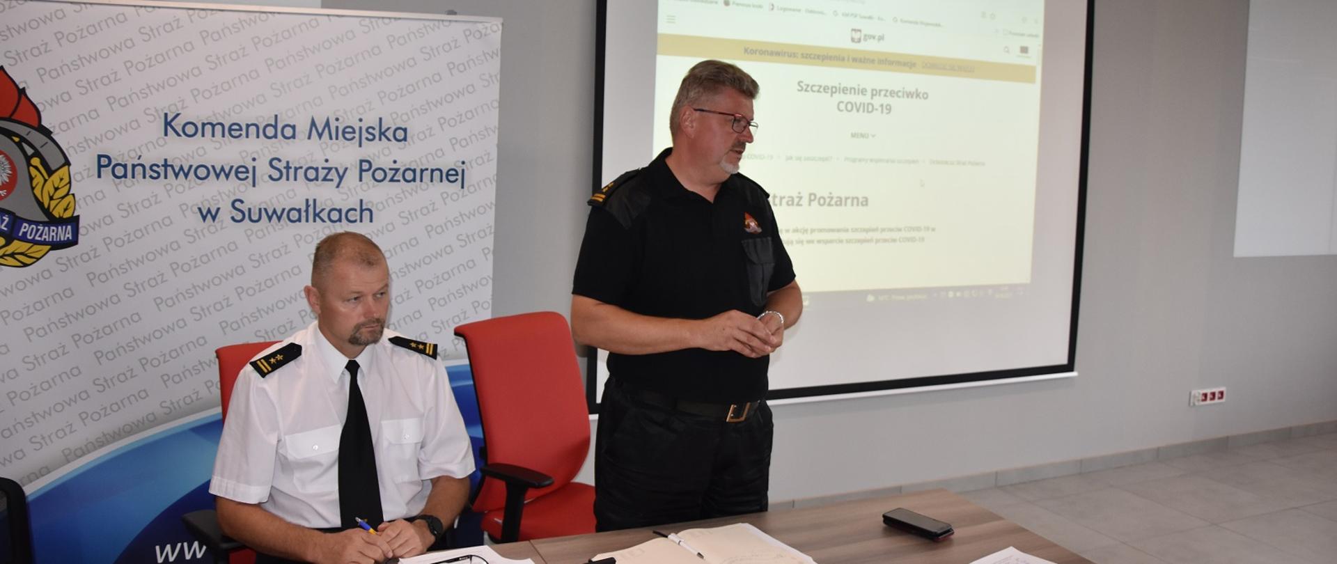 Zdjęcie obrazuje 2 strażaków, jeden stoi i referuje. W tle slajd o treści szczepienie przeciw COVID-19 .