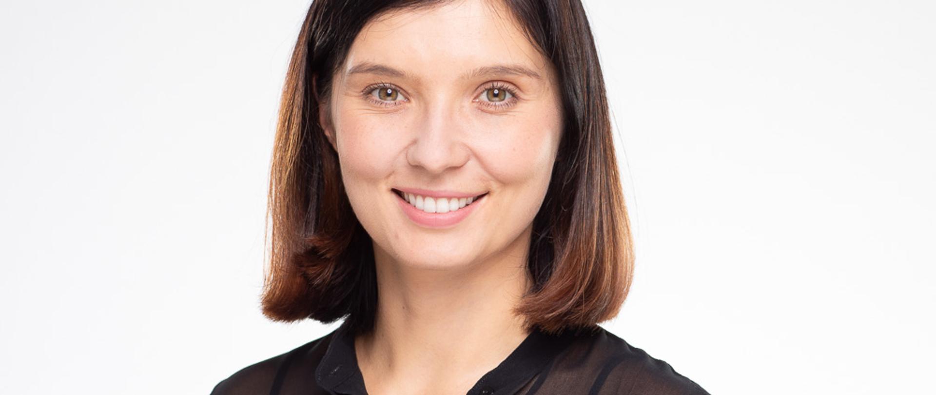 Młoda około czterdziestoletnia kobieta uśmiechnięta, ma ciemne oczy oraz krótkie do ramion włosy w kolorze brąz. Ubrana w czarną elegancką koszulę. 